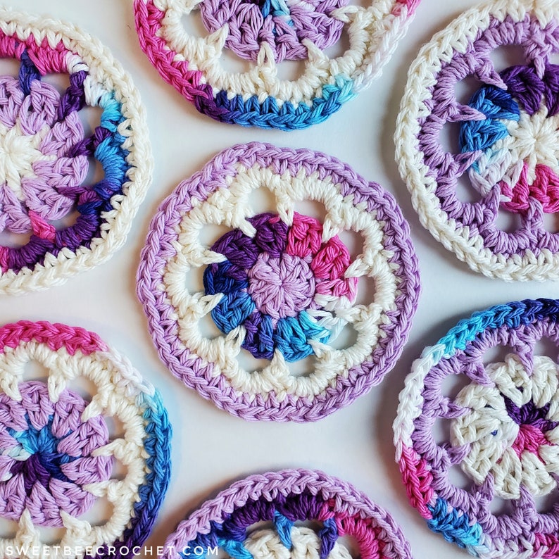 K&ouml;nnte beinhalten: Sechs geh&auml;kelte Untersetzer in den Farben Lila, Rosa und Blau. Die Untersetzer haben eine wei&szlig;e Mitte mit einem kreisf&ouml;rmigen Design.