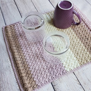 May include: A crocheted trivet with stripes of beige, light pink, and white. Two clear glass cups and a purple mug are sitting on the trivet. The trivet has the text "sweetbecrochet.com" on the side.