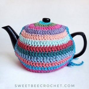 Può includere: Un copriteiera in crochet colorato con un motivo a righe in tonalità di rosa, blu, verde e rosso. Il copriteiera è realizzato con punto basso e ha un lungo anello in crochet per appenderlo.