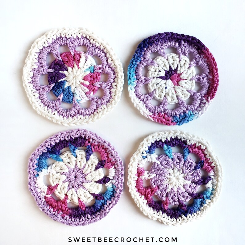 K&ouml;nnte beinhalten: Vier geh&auml;kelte Untersetzer in den Farben Lila, Rosa und Blau. Jeder Untersetzer hat eine wei&szlig;e Mitte mit einem Blumenmuster.