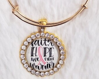 May include: Gold bangle bracelet with a charm featuring a pink ribbon, rhinestones, and the words "faith hope love fight warrior".