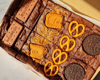 May include: A box of assorted brownie squares with different toppings. The brownies are topped with Oreo cookies, pretzels, and Bourbon biscuits. Some of the brownies have a swirl of caramel.