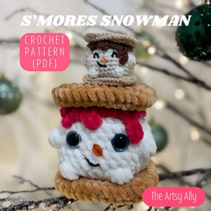 May include: A crocheted s'mores snowman ornament with a white marshmallow body, pink frosting, and brown graham cracker cookies. The snowman has googly eyes and a carrot nose. The ornament is hanging from a branch.