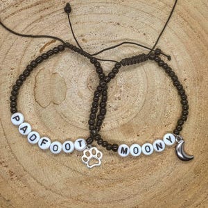 May include: Two brown beaded bracelets with white beads spelling out the names "Padfoot" and "Moony". One bracelet has a silver paw print charm, and the other has a silver crescent moon charm.