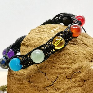 Puede incluir: Un brazalete de cordón negro con siete cuentas de piedras preciosas de colores. Las cuentas son moradas, azules, verdes, amarillas, naranjas, rojas y negras.