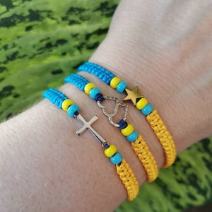 May include: Three friendship bracelets with yellow and blue braided cords and beads. The bracelets have charms including a cross, a heart, and a star.