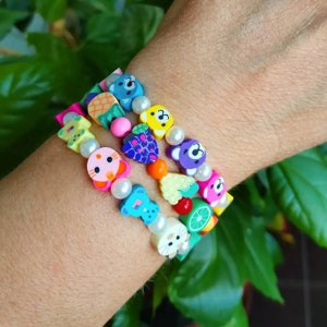 Peut inclure: Trois bracelets colorés avec différents charms, dont des fruits, des animaux et des cœurs. Les bracelets sont faits de perles en plastique et de perles blanches.