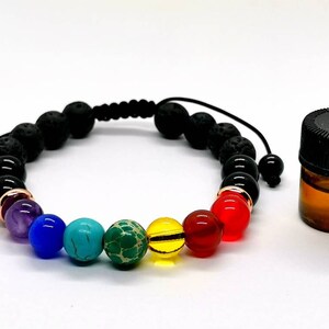 Op de afbeelding: Een zwarte lavastenen armband met regenboogkleurige kralen en een klein flesje etherische olie. De armband heeft een zwart koord met een schuifknoop om de maat aan te passen.