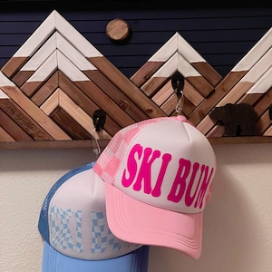 May include: Two trucker hats hang from a wooden mountain-themed wall decor. One hat is pink with "SKI BUM" in bold letters, the other is blue with a checkered pattern. The wall decor features a mountain range design.