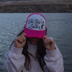 May include: A pink and white trucker hat with the text "Ski Ya Later" in black script. The hat features a skier graphic and is being held up by a person.