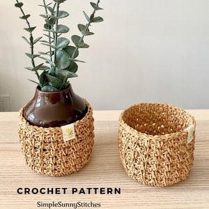 May include: Two crocheted plant pot covers in a natural beige color. The covers are made with a textured stitch pattern. The covers are sitting on a wooden surface. The text "CROCHET PATTERN" and "SimpleSunnyStitches" is visible below the covers.