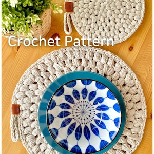 May include: Two beige crochet pot holders with a brown leather loop. The larger pot holder is on a wooden table with a blue and white patterned bowl on top.