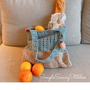 May include: A crocheted tote bag with a blue and beige color scheme. The bag is filled with a loaf of bread and oranges. The bag has a small crocheted charm attached to the handle. The text "Simple Sunny Stitches" is visible in the bottom right corner of the image.