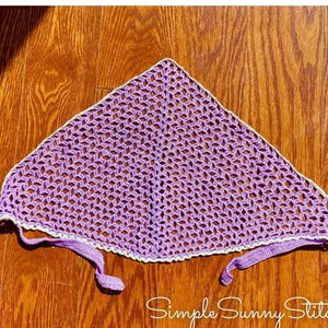 May include: A purple crochet head scarf with a white crocheted border. The scarf has two long ties that can be tied at the back of the neck.  Simple Sunny Stitches is written in white text on a brown wooden surface.