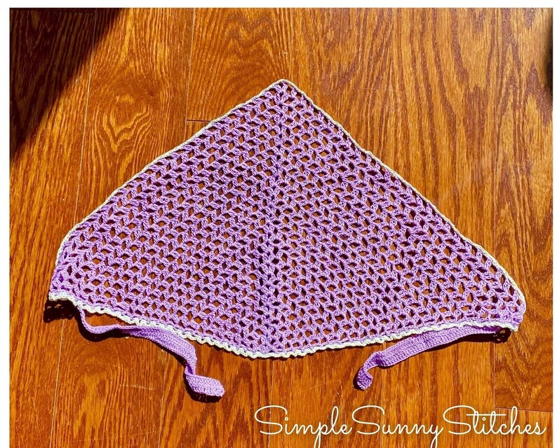 May include: A purple crochet head scarf with a white crocheted border. The scarf has two long ties that can be tied at the back of the neck.  Simple Sunny Stitches is written in white text on a brown wooden surface.