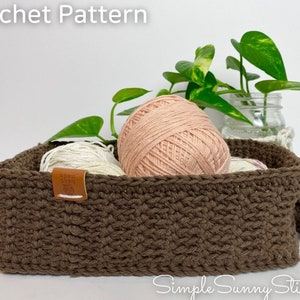 Op de afbeelding: Een bruine gehaakte mand met twee bollen garen erin. De mand heeft twee handvatten en staat op een wit oppervlak. Achter de mand staat een plant in een pot met groene bladeren.