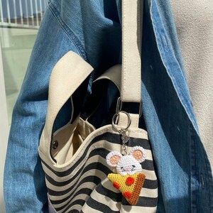 May include: A white crocheted mouse holding a slice of pizza with red pepperoni, attached to a black and white striped tote bag with a beige strap. The bag has a small pocket on the side. The text "Simple Sunny Stitches" is visible at the bottom of the image.
