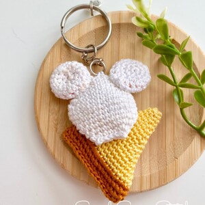 May include: White crocheted ice cream cone keychain with a yellow and orange cone and a white crocheted ice cream scoop with Mickey Mouse ears. The keychain is on a wooden surface with a green plant in the background.