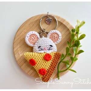May include: A white crocheted mouse holding a crocheted pizza slice keyring with red and yellow accents. The keyring has a silver charm that says "Made with love".
