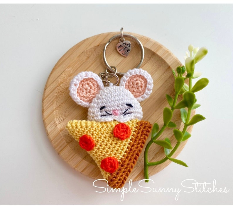May include: A white crocheted mouse holding a crocheted pizza slice keyring with red and yellow accents. The keyring has a silver charm that says "Made with love".