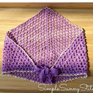 May include: A purple crocheted shawl with a white crocheted border and a bow tie. The shawl is laid flat on a wooden surface. The text "Simple Sunny Stitches" is visible in the bottom right corner of the image.
