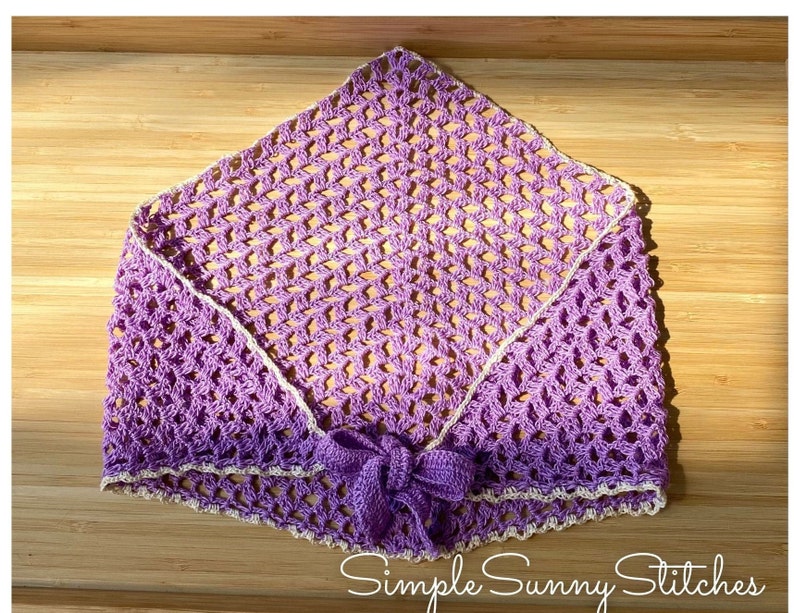 May include: A purple crocheted shawl with a white crocheted border and a bow tie. The shawl is laid flat on a wooden surface. The text "Simple Sunny Stitches" is visible in the bottom right corner of the image.