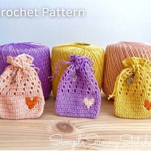 May include: Three crocheted drawstring bags in pink, purple, and yellow, with a heart design. The bags are sitting on a wooden surface with balls of yarn in the background. The text "Crochet Pattern" is at the top of the image. The text "Simple Sunny Stitches" is at the bottom of the image.