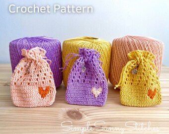 May include: Three crocheted drawstring bags in pink, purple, and yellow, with a heart design. The bags are sitting on a wooden surface with balls of yarn in the background. The text "Crochet Pattern" is at the top of the image. The text "Simple Sunny Stitches" is at the bottom of the image.