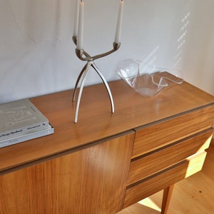 Op de afbeelding: Een houten dressoir met drie lades en een zilveren kandelaar met drie brandende kaarsen. Het dressoir staat op een houten vloer en er staat een glazen kom op de bovenkant van het dressoir.