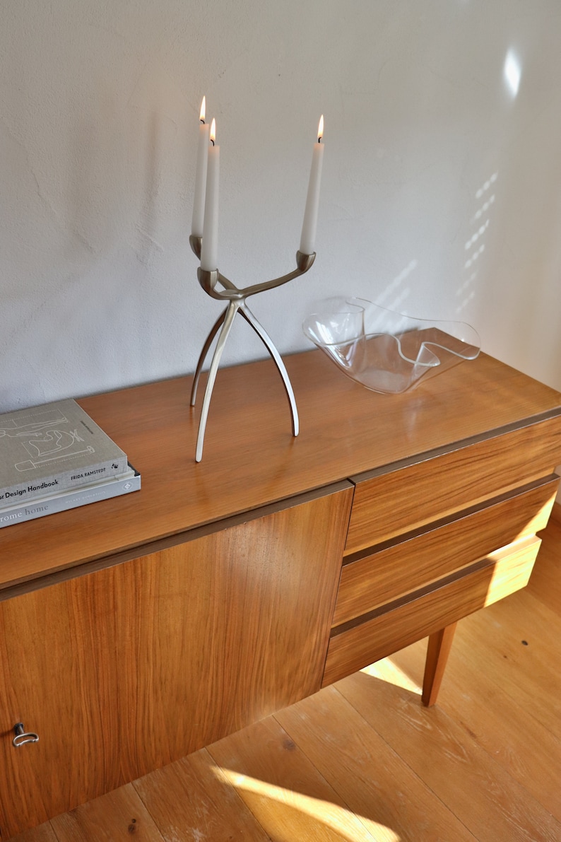 Op de afbeelding: Een houten dressoir met drie lades en een zilveren kandelaar met drie brandende kaarsen. Het dressoir staat op een houten vloer en er staat een glazen kom op de bovenkant van het dressoir.