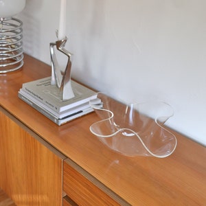 Op de afbeelding: Een houten dressoir met twee laden en een transparante plastic kom erop. Een zilveren kandelaar met een witte kaars staat op het dressoir naast een stapel boeken.