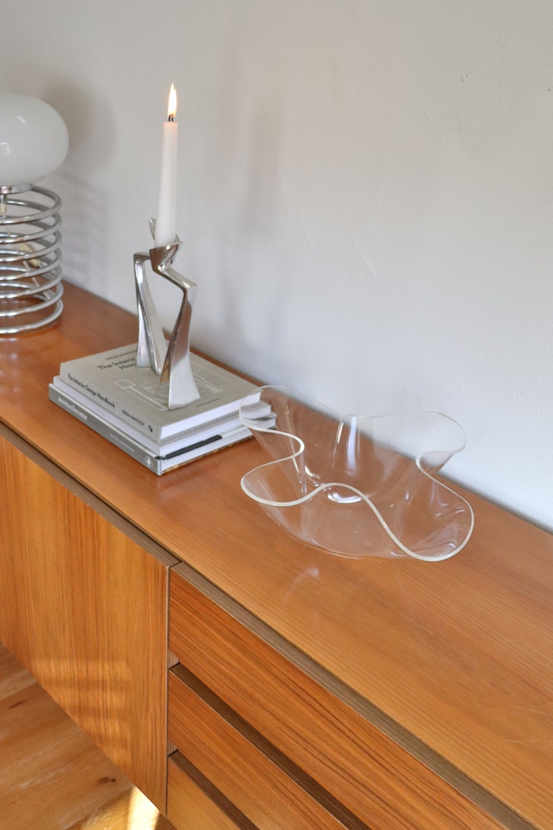 Op de afbeelding: Een houten dressoir met twee laden en een transparante plastic kom erop. Een zilveren kandelaar met een witte kaars staat op het dressoir naast een stapel boeken.