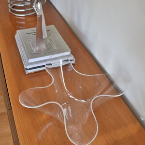 Op de afbeelding: Een transparante plastic kom met een uniek, golvend ontwerp staat op een houten oppervlak. De kom is leeg en lijkt van een duurzaam materiaal te zijn gemaakt.