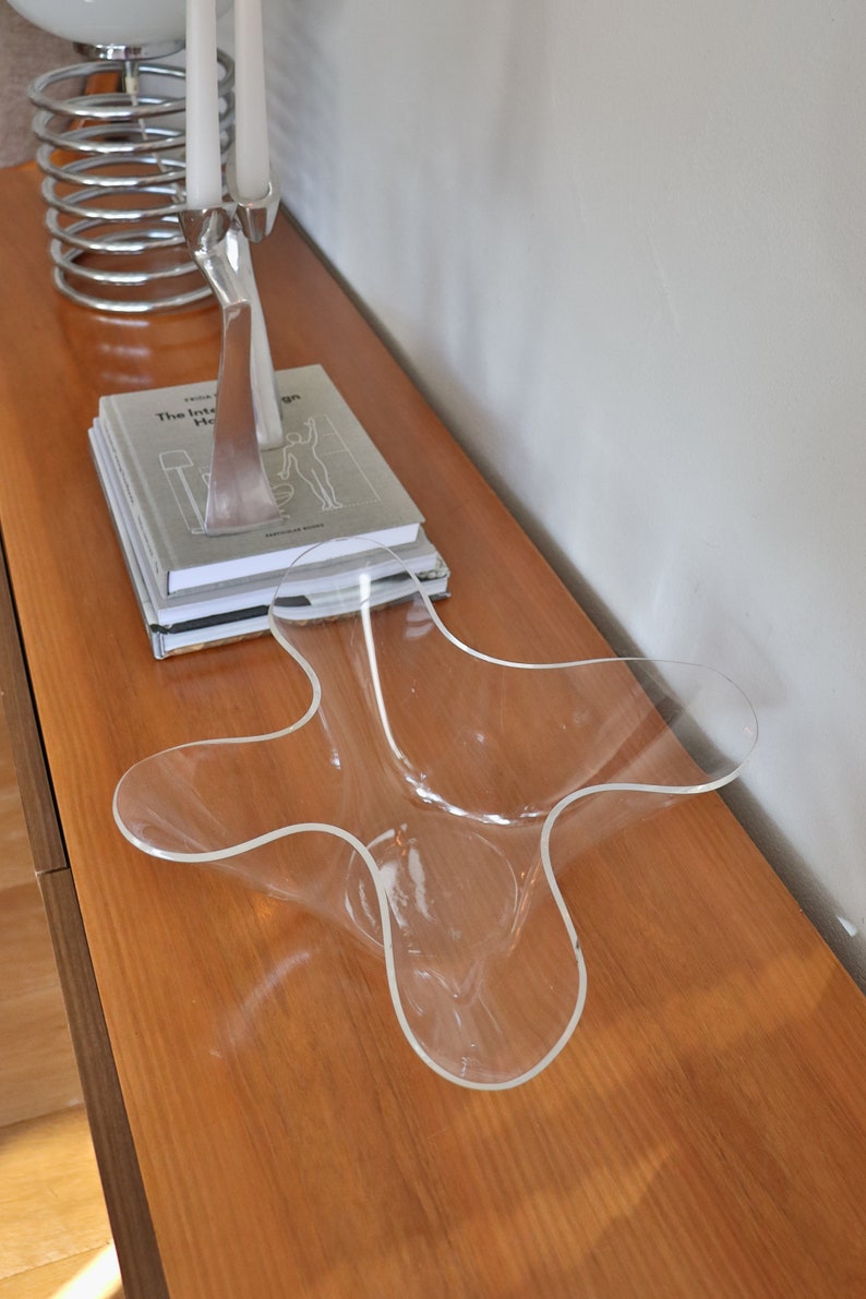 Op de afbeelding: Een transparante plastic kom met een uniek, golvend ontwerp staat op een houten oppervlak. De kom is leeg en lijkt van een duurzaam materiaal te zijn gemaakt.
