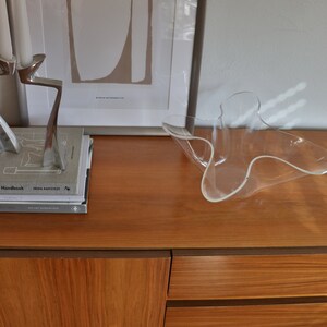 Op de afbeelding: Een houten kast met twee lades en een deur. De kast is gemaakt van lichtbruin hout en heeft een donkerbruine rand. Een glazen kom staat op de kast. De kast staat voor een witte muur. Er hangt een ingelijst printje op de kast met de tekst "THE ABSTRACT ART COLLECTION" en "MIRROR AUTOMATICS".