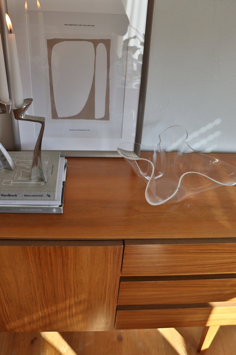 Op de afbeelding: Een houten kast met twee lades en een deur. De kast is gemaakt van lichtbruin hout en heeft een donkerbruine rand. Een glazen kom staat op de kast. De kast staat voor een witte muur. Er hangt een ingelijst printje op de kast met de tekst "THE ABSTRACT ART COLLECTION" en "MIRROR AUTOMATICS".