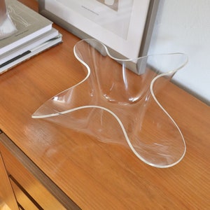 Op de afbeelding: Een heldere acryl kom met een uniek, golvend, stervormig ontwerp. De kom staat op een houten oppervlak.