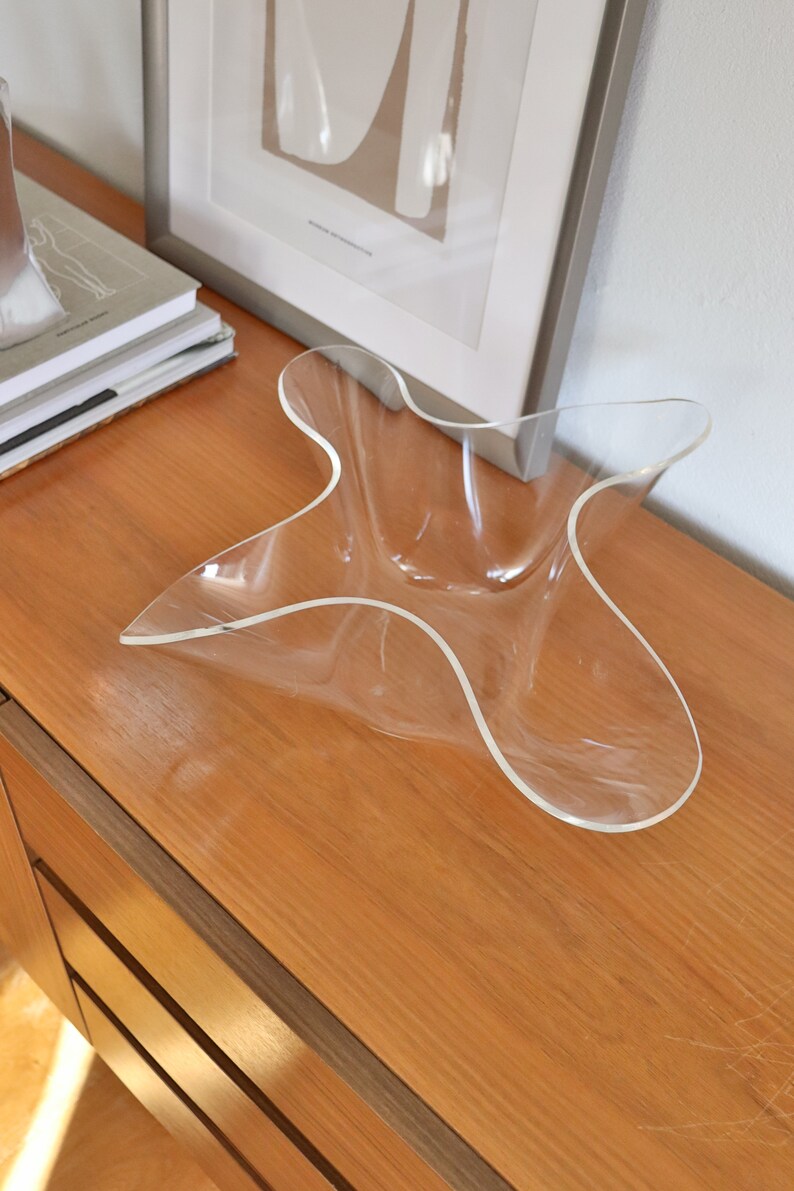 Op de afbeelding: Een heldere acryl kom met een uniek, golvend, stervormig ontwerp. De kom staat op een houten oppervlak.