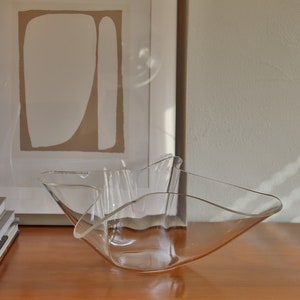 Op de afbeelding: Een heldere glazen kom met een golvende, organische vorm staat op een houten oppervlak. De kom is leeg en lijkt van dun glas te zijn gemaakt.