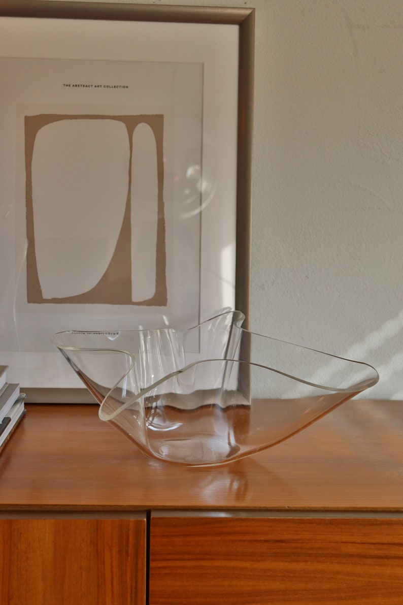 Op de afbeelding: Een heldere glazen kom met een golvende, organische vorm staat op een houten oppervlak. De kom is leeg en lijkt van dun glas te zijn gemaakt.
