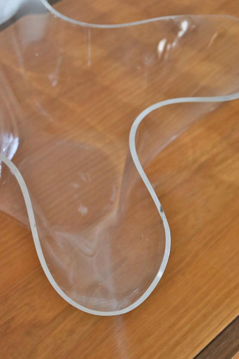 Op de afbeelding: Een doorzichtige plastic kom met een unieke, golvende, organische vorm. De kom staat op een houten oppervlak.