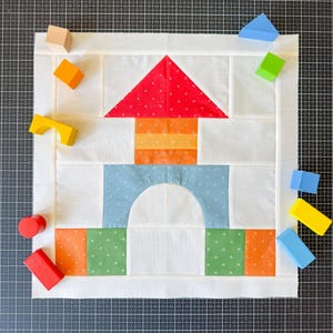 May include: A white fabric quilt with a red, orange, yellow, blue, and green house design. The quilt is surrounded by wooden blocks in various colors.