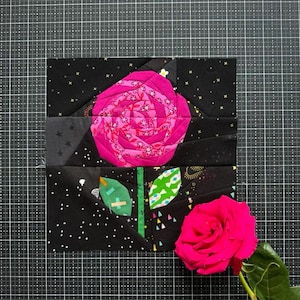 May include: A square fabric quilt block featuring a stylized rose design in shades of hot pink and magenta, set against a black background with white star patterns. A green stem and leaves are visible, accompanied by a fresh hot pink rose flower.
