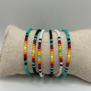 May include: Six beaded bracelets in various colours, including turquoise, black, white, red, yellow, and orange. The bracelets are displayed on a beige fabric surface, showcasing their colourful patterns and designs. These bracelets are perfect for adding a pop of colour to any outfit.