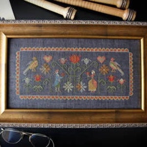 May include: A framed cross-stitch embroidery depicting a whimsical garden scene with two figures, birds, and flowers. The embroidery is stitched on a dark blue fabric and framed in a gold-colored frame.