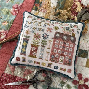 May include: A decorative, square pillow with a cross-stitch design featuring a house, spools, and the text "button house." The pillow has a teal border and is on a floral quilt. A pair of ornate scissors rests nearby.