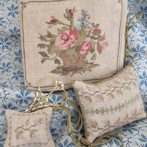 May include: A square linen piece featuring a cross-stitched floral basket, two rectangular linen pincushions with embroidered floral borders, and gold-toned metal embroidery scissors with an intricate handle design and attached green cord.