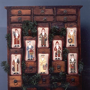 May include: A vintage wooden cabinet with twelve drawers, each containing a hand-stitched cross-stitch design of a different Santa Claus figure. The Santa Claus figures are dressed in various colors and styles, including red, green, blue, and brown.