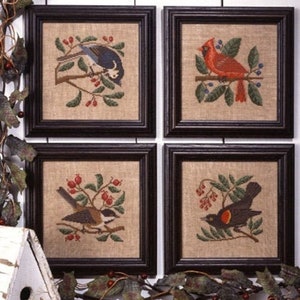 May include: Four framed cross-stitch artworks featuring birds on branches with berries. The birds are depicted in various colors, including blue, red, brown, and black. The artwork is framed in dark brown frames and set against a neutral background.