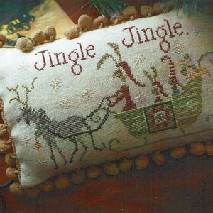 May include: A white pillow with a cross-stitch design of a reindeer pulling a sleigh with the text "Jingle Jingle" in red. The pillow is trimmed with brown pom-poms.